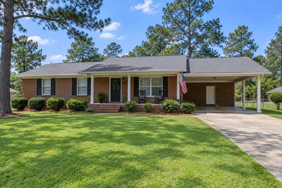Single-story brick ranch house with shuttered windows and a covered carport, an American flag on a post, a green lawn and tall pine trees under a blue sky