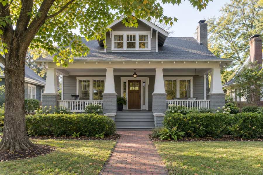 Grey Craftsman bungalow with a wide covered porch, tapered columns, white trim, a front dormer, a brick walk, a mature maple and a low hedge in soft morning light