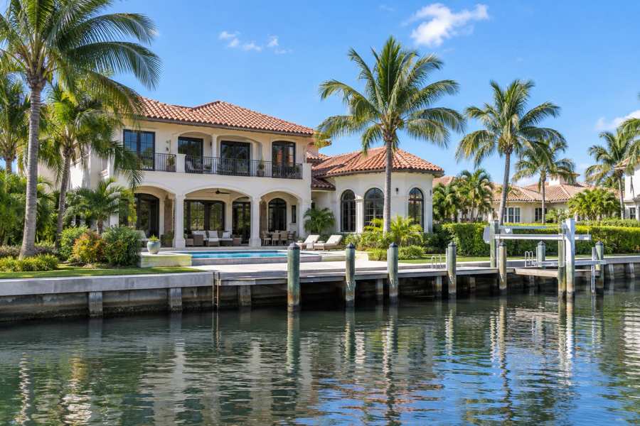 Modern Mediterranean waterfront house with a terracotta barrel tile roof, palm trees, a pool and a private dock on a calm canal in bright afternoon light
