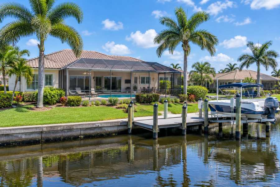 Single-story stucco house with a tile roof, a screened pool enclosure and a boat on a lift at a wooden dock on a canal, with tall palm trees, green lawn and a bright blue sky
