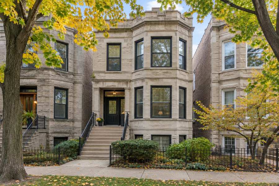 Two-story limestone Chicago greystone with bay windows, a front stoop with black iron railings and a black iron fence, on a tree-lined street with yellow autumn maple leaves