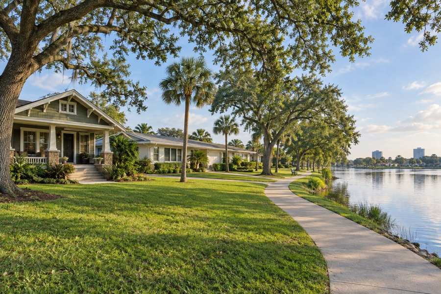 Green craftsman bungalow with a covered porch and stone piers beside a cream ranch house, on a lawn beside a paved lakeside walking path, with live oaks, palms and a small lake under a blue sky