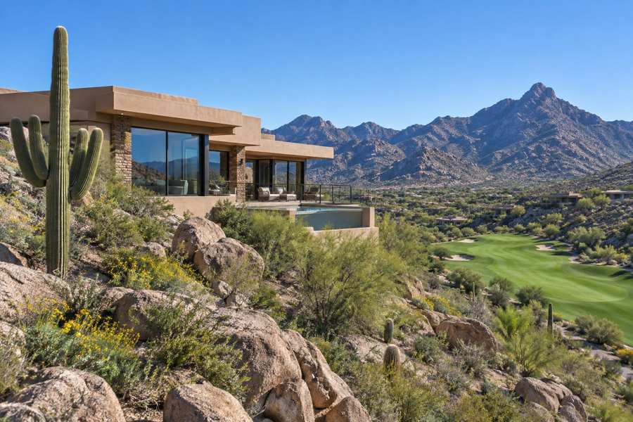 Contemporary desert home with sand-colored stucco walls and large glass windows on a rocky hillside with saguaro cactus, a green golf fairway below and mountain ridges beyond