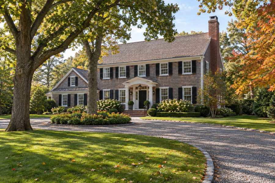 Two story cedar shingle colonial house with white trim, black shutters, a columned entry portico and a brick chimney, set behind a gravel circular driveway and a manicured lawn under large oak and maple trees in early autumn