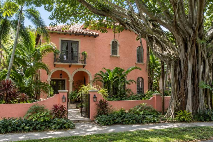 Two story 1920s Mediterranean Revival house with coral pink stucco walls, arched openings and a clay tile roof, shaded by a large banyan tree and tropical plants