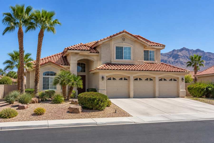 Two-story beige stucco house with a red clay tile roof, a three-car garage, palm trees and desert landscaping on a quiet street, with a mountain ridge in the distance under a clear blue sky