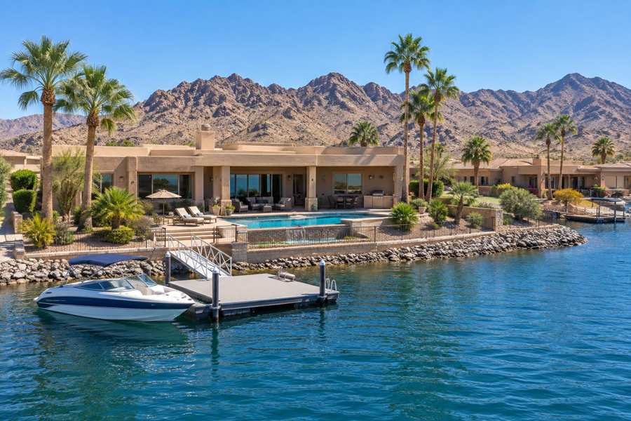 Southwestern stucco lakefront house with a flat roof and a pool, a boat dock with a speedboat on blue water, palm trees and tan desert mountains under a clear sky