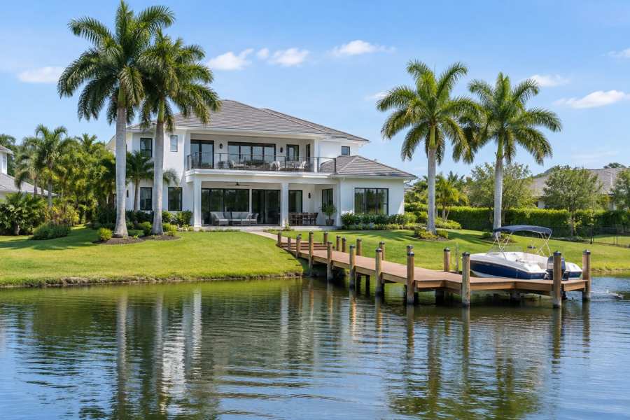 Large white two-story contemporary lakefront house with a covered terrace and a long wooden dock with a small boat on calm water, a lawn running to the lake and royal palms