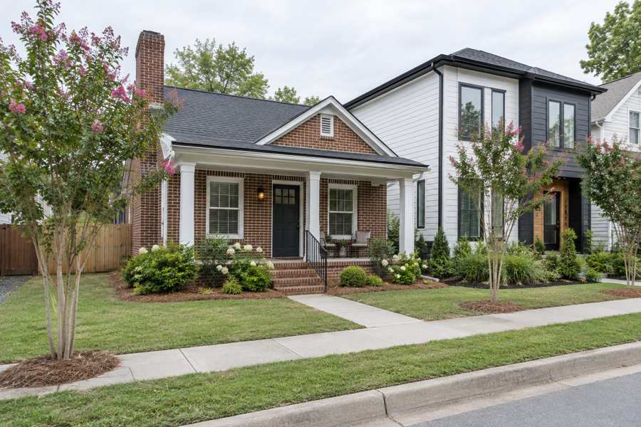 Small red brick cottage with a covered porch, white columns and a dark front door beside a new white and black two-storey house, with pink flowering crepe myrtles, a sidewalk and lawn under a grey sky