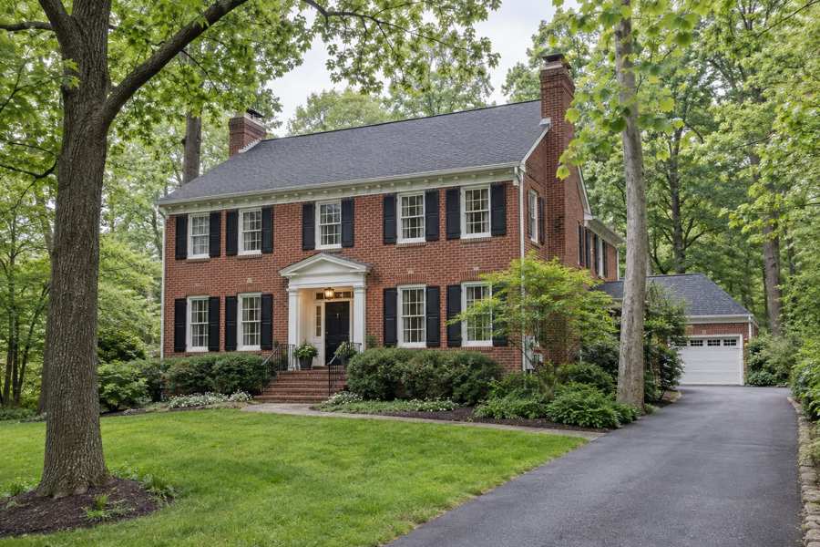 Illustrative red brick Georgian-style house with white trim, black shutters, a paved driveway and tall trees on a wooded lawn.