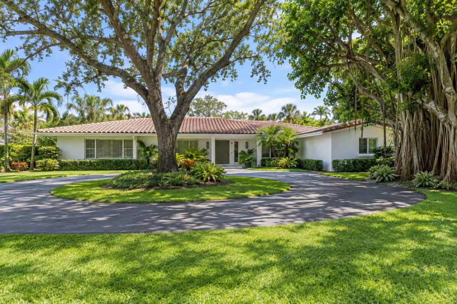 White stucco 1960s ranch style house with a low barrel tile roof, a circular driveway and mature oak and banyan trees on a green lawn