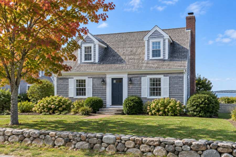 Illustrative gray shingled house with two dormers, white trim, a dark front door, brick chimney, flowering shrubs, autumn tree and a low stone wall.