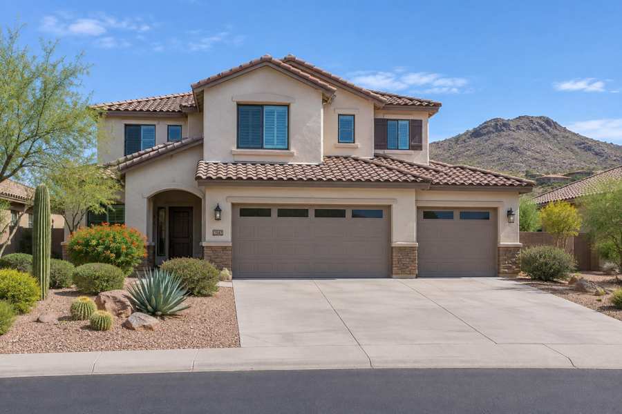 Two-story beige stucco house with a clay tile roof, a three-car garage with brown doors, a small arched entry and desert landscaping with cactus and agave, with a rocky hill behind under a blue sky