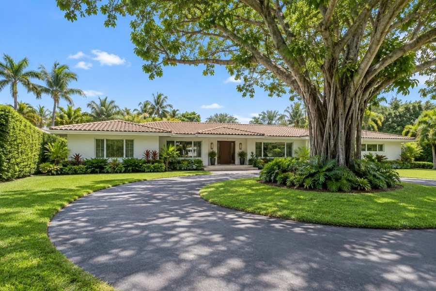 Single-story white stucco ranch house with a low tile roof and a circular driveway on a large green lot, with a mature banyan tree and a tall tropical hedge under a blue sky