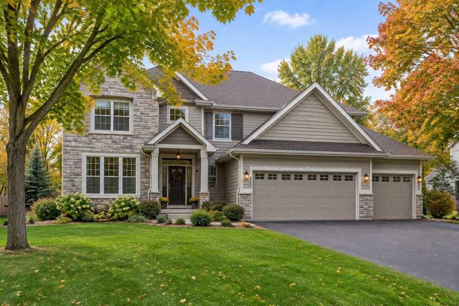 Two-story suburban house with stone and tan siding, a covered front entry, a three-car garage and a dark asphalt driveway, with a green lawn and maple trees in yellow and orange autumn color