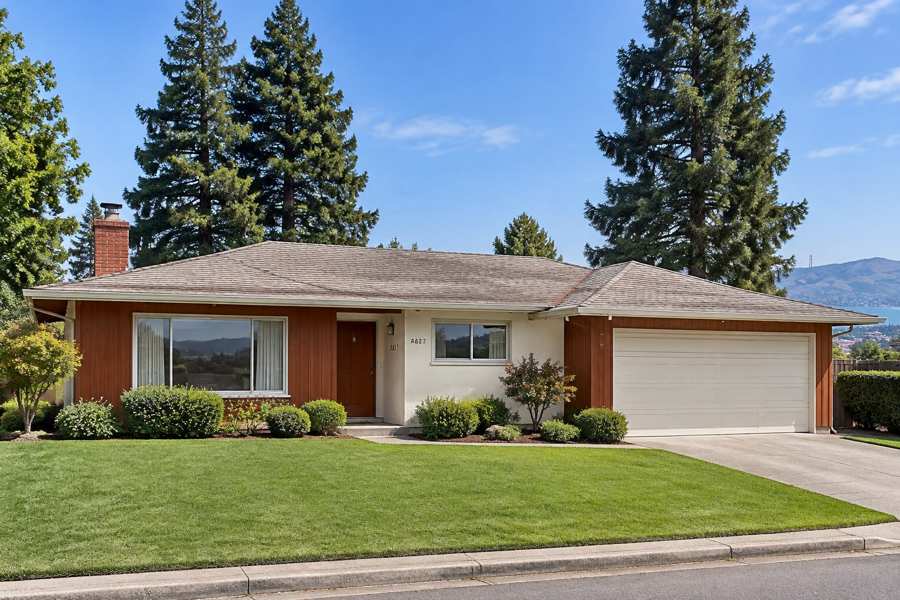 Single-story ranch house with red-brown vertical wood siding and cream stucco, a wide picture window, a brick chimney, a cream two-car garage door and a neat lawn, with tall evergreen trees and distant hills and water under a blue sky
