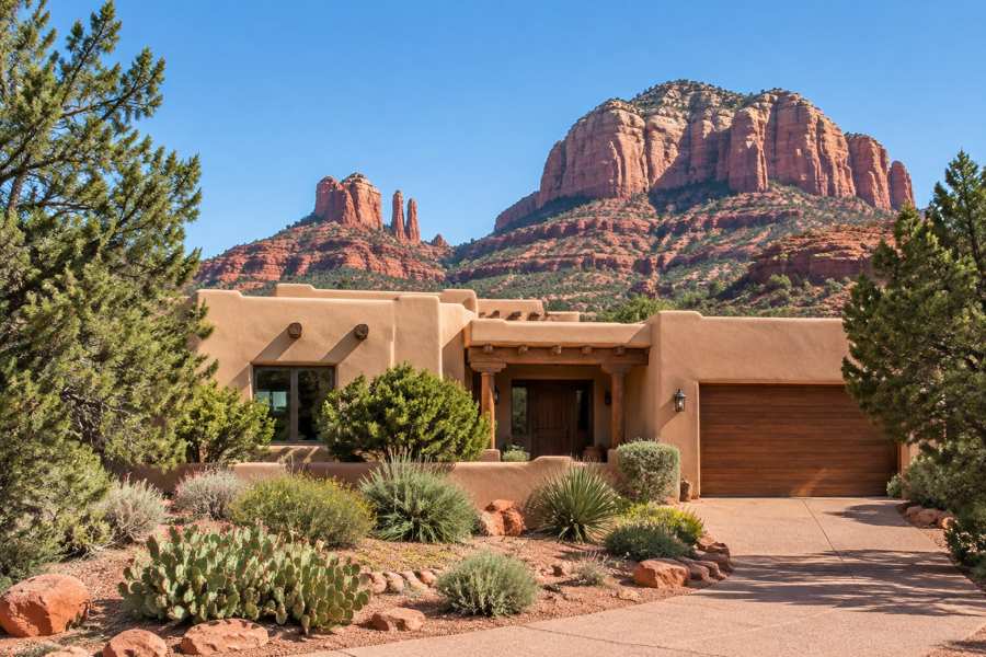 Adobe-style tan stucco home with a timber-framed entry and a wooden garage door, desert shrubs, prickly pear cactus and juniper trees in front, and red rock buttes rising behind under a clear blue sky