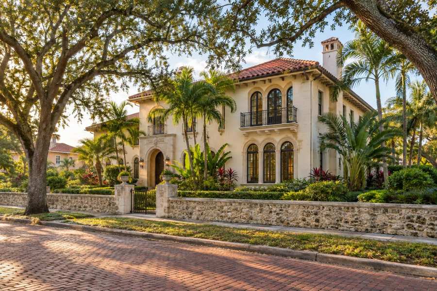 Cream stucco Mediterranean Revival house with a barrel tile roof, arched windows and an iron balcony behind a coquina stone wall, with live oaks, palms and tropical plants beside a brick paved street in golden light