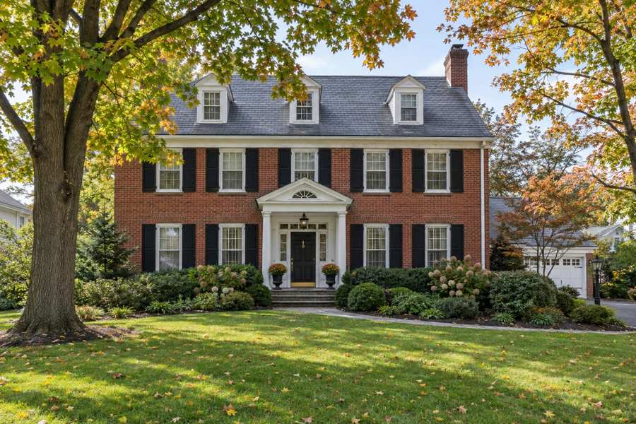 Red brick Colonial house with black shutters, white trim, a columned front porch with a black door and a slate roof with three dormers, set behind a green lawn and large maple trees with orange autumn leaves