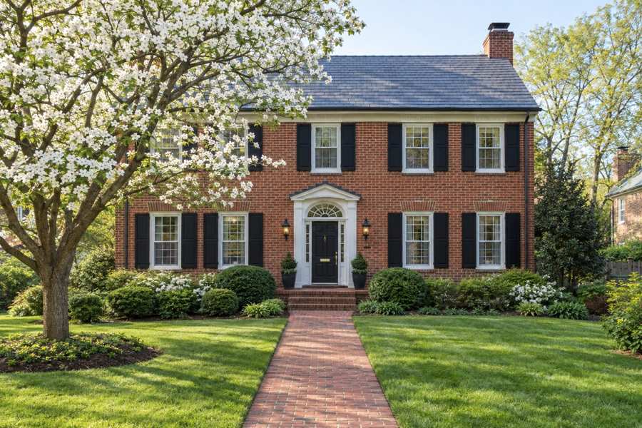 Brick center-hall Colonial house with black shutters and a slate roof, a dogwood tree in bloom, a manicured lawn and a brick walkway in soft morning light
