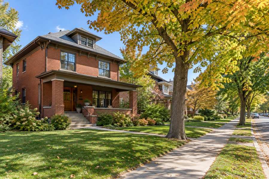 Illustrative red brick two-story house with a covered front porch, green lawn, sidewalk and a large maple with golden autumn leaves.