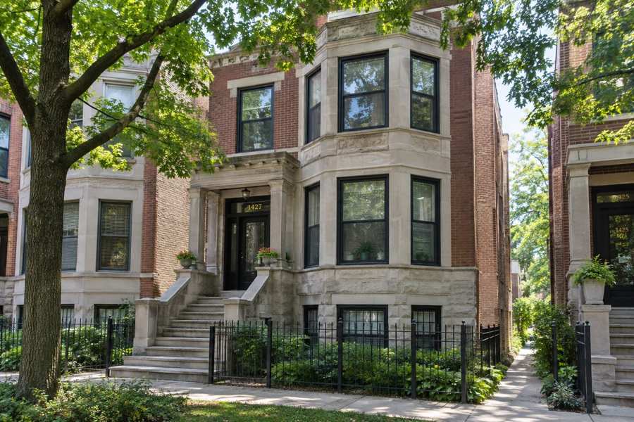 Red brick and limestone multi-unit building with a two-story stone bay window, a columned stone entry with a staircase and a black iron fence, on a tree-lined sidewalk with green leaves in summer